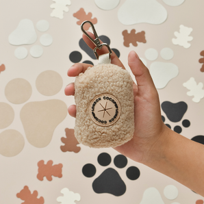 Hand met de crèmekleurige Teddy Rupert poepzakhouder, met zichtbare clip en ronde patch