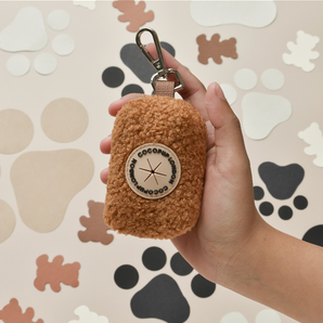 Hand holding the brown Teddy Paddington poop bag holder with clip and round patch visible