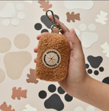 Hand met de bruine Teddy Paddington poepzakhouder, met zichtbare clip en ronde patch